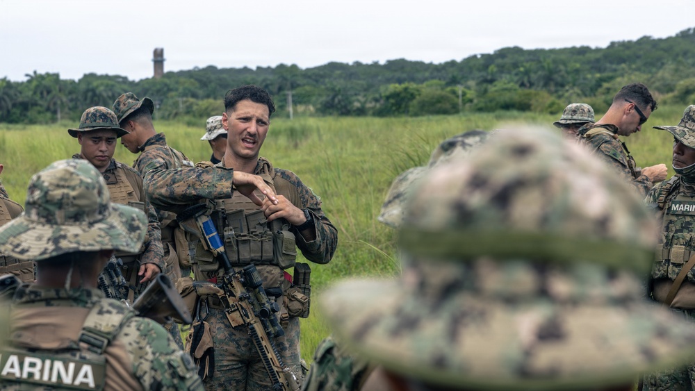 The image shows a group of military personnel in a field setting, appearing to be engaged in a briefing or training exercise. A central figure in US Marine Corps MARPAT camouflage is gesturing and speaking to the assembled group. The presence of 'MARINA' text on one uniform suggests this may be a joint exercise involving Mexican Naval Infantry (Infantería de Marina) and US Marines.