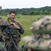 The image shows a group of military personnel in a field setting, appearing to be engaged in a briefing or training exercise. A central figure in US Marine Corps MARPAT camouflage is gesturing and speaking to the assembled group. The presence of 'MARINA' text on one uniform suggests this may be a joint exercise involving Mexican Naval Infantry (Infantería de Marina) and US Marines.