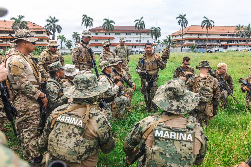 A multinational military briefing or training exercise is taking place outdoors on a grassy field. Personnel wearing various camouflage uniforms, including those marked 'MARINA' (Mexican Navy), are gathered in a group while one individual appears to be addressing the group. The setting includes tropical palm trees and multi-story buildings in the background, suggesting a Latin American or Caribbean location.
