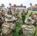A multinational military briefing or training exercise is taking place outdoors on a grassy field. Personnel wearing various camouflage uniforms, including those marked 'MARINA' (Mexican Navy), are gathered in a group while one individual appears to be addressing the group. The setting includes tropical palm trees and multi-story buildings in the background, suggesting a Latin American or Caribbean location.