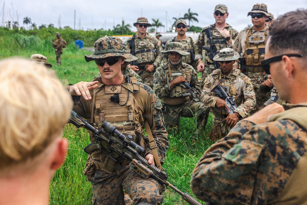A group of military personnel in mixed camouflage uniforms are gathered in an outdoor briefing or debriefing session in what appears to be a tropical environment. A central figure wearing a boonie hat and plate carrier vest, holding an assault rifle, appears to be addressing the group. Multiple nationalities appear to be represented, as indicated by varying uniform patterns and at least one 'MARINA' patch visible on a uniform.