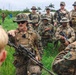 A group of military personnel in mixed camouflage uniforms are gathered in an outdoor briefing or debriefing session in what appears to be a tropical environment. A central figure wearing a boonie hat and plate carrier vest, holding an assault rifle, appears to be addressing the group. Multiple nationalities appear to be represented, as indicated by varying uniform patterns and at least one 'MARINA' patch visible on a uniform.
