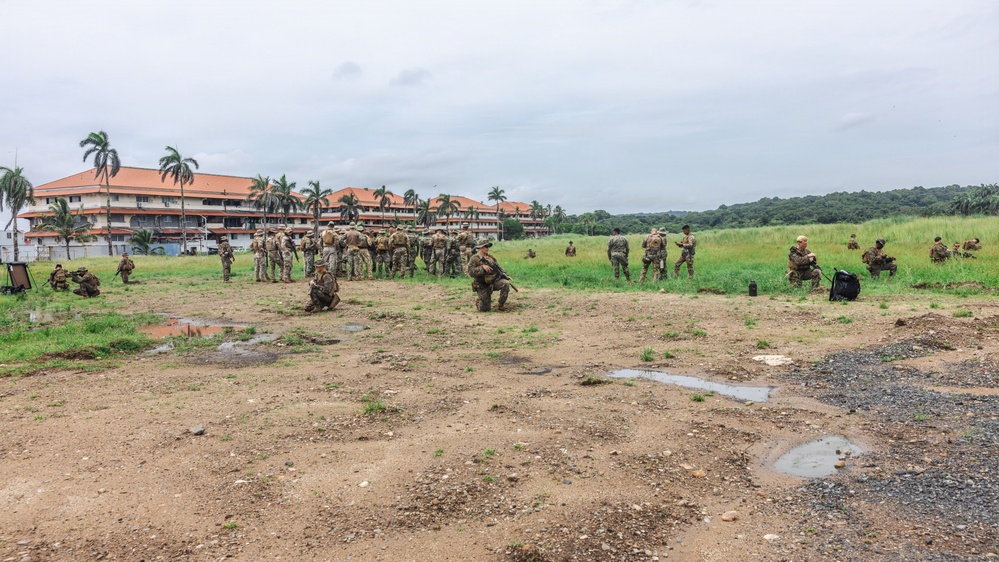 A large group of military personnel in camouflage uniforms are gathered on an open field during what appears to be a training exercise or briefing. The setting includes a multi-story building with an orange/terracotta roof surrounded by palm trees, suggesting a tropical or subtropical location. Some soldiers are kneeling in tactical positions while others are standing in a clustered group.