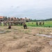 A large group of military personnel in camouflage uniforms are gathered on an open field during what appears to be a training exercise or briefing. The setting includes a multi-story building with an orange/terracotta roof surrounded by palm trees, suggesting a tropical or subtropical location. Some soldiers are kneeling in tactical positions while others are standing in a clustered group.