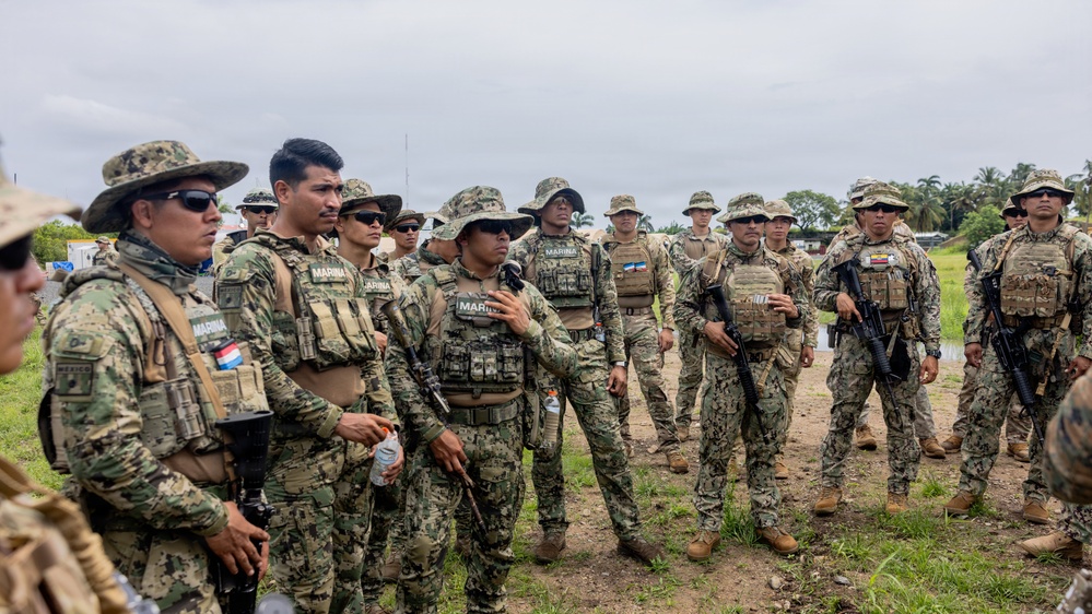 A group of military personnel in camouflage uniforms are gathered outdoors in what appears to be a briefing or assembly. Multiple patches reading 'MARINA' are visible, indicating Mexican Naval Infantry (Infantería de Marina), along with a Mexican flag patch on at least one uniform. Personnel are equipped with tactical vests, weapons, and various headgear including boonie hats and baseball caps.