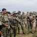 A group of military personnel in camouflage uniforms are gathered outdoors in what appears to be a briefing or assembly. Multiple patches reading 'MARINA' are visible, indicating Mexican Naval Infantry (Infantería de Marina), along with a Mexican flag patch on at least one uniform. Personnel are equipped with tactical vests, weapons, and various headgear including boonie hats and baseball caps.