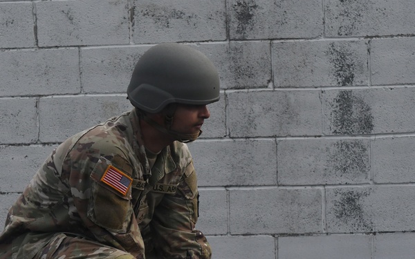A U.S. Army soldier in full combat uniform is crouching on an elevated platform or ledge against a concrete block wall. The soldier is wearing OCP (Operational Camouflage Pattern) uniform, a ballistic helmet, and black tactical gloves. The posture suggests a tactical or training exercise scenario.
