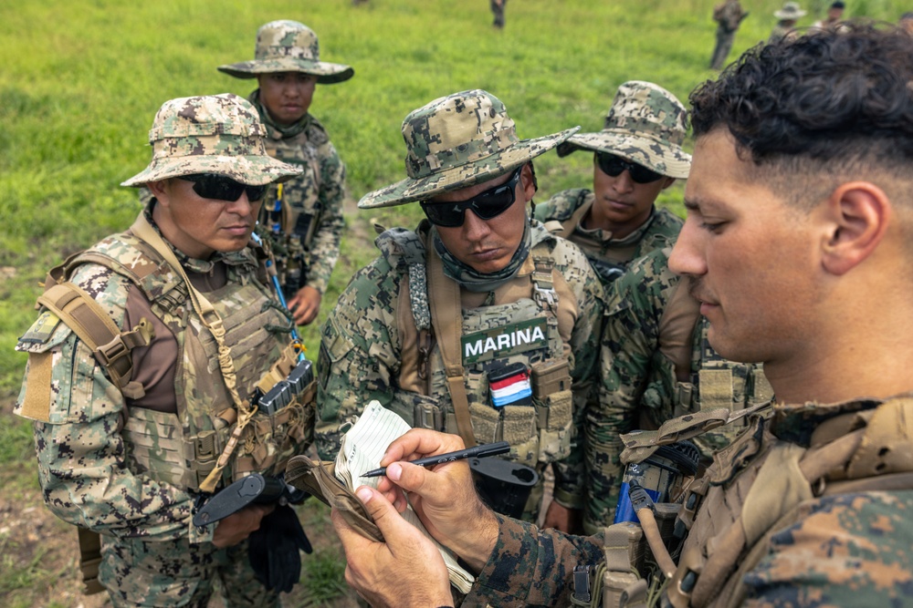 A group of military personnel in camouflage uniforms are gathered in a field, appearing to review notes or receive a briefing. One soldier wearing a 'MARINA' patch with what appears to be a Paraguayan flag is prominently visible in the center. A soldier on the right is writing in a small notepad while others observe.