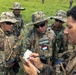 A group of military personnel in camouflage uniforms are gathered in a field, appearing to review notes or receive a briefing. One soldier wearing a 'MARINA' patch with what appears to be a Paraguayan flag is prominently visible in the center. A soldier on the right is writing in a small notepad while others observe.