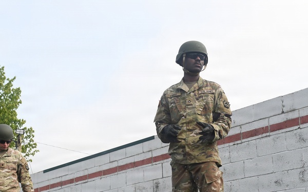 Two U.S. Army soldiers in full combat uniforms, including OCP camouflage, helmets, gloves, and tan boots, are standing on an elevated platform or beam at what appears to be a military training obstacle course. The soldier in the foreground faces forward confidently while the second soldier looks downward. A white cinder block wall with a red stripe is visible in the background.