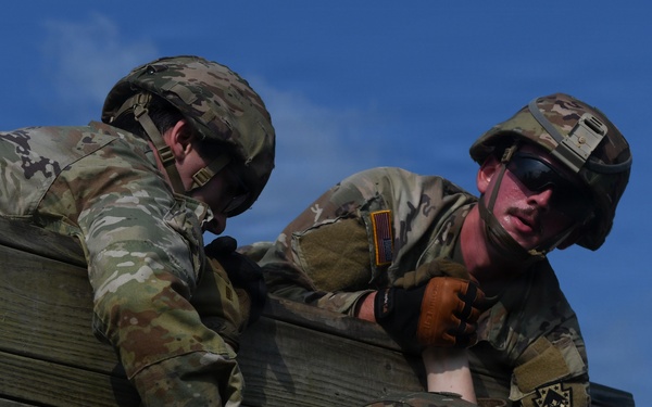 Two soldiers in US Army Operational Camouflage Pattern (OCP) uniforms are navigating a wooden wall obstacle, with one soldier assisting the other over the barrier. Both wear tactical helmets and combat gear, suggesting this is a military training exercise or obstacle course. The image is taken from a low angle against a clear blue sky.