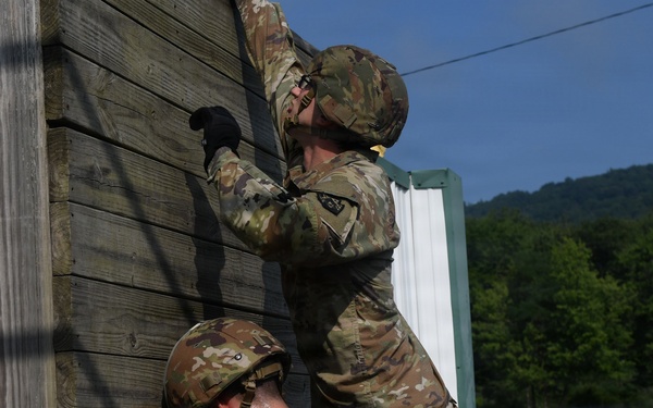 Two soldiers in US Army OCP (Operational Camouflage Pattern) uniforms and helmets are scaling or climbing a wooden log wall obstacle, consistent with a military training exercise or obstacle course. One soldier is higher on the wall while the other appears to be providing a boost or preparing to follow. The setting appears to be a military training facility surrounded by green hills and open fields.