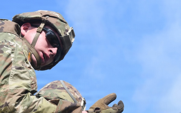 A soldier in full military combat uniform is leaning over a wooden structure, appearing to put on or adjust tactical gloves. The soldier is wearing OCP (Operational Camouflage Pattern) uniform, a combat helmet with sunglasses flipped up, and tan tactical gloves. The image is taken from a low angle against a partly cloudy blue sky.