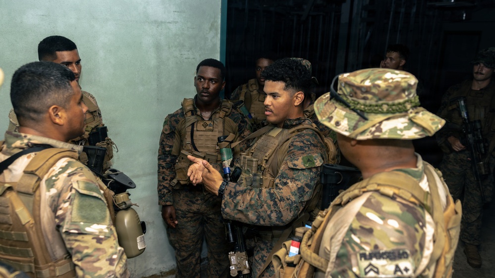A group of military personnel in two distinct camouflage uniforms are gathered in what appears to be a briefing or coordination session inside a building. Some wear MARPAT (Marine pattern) uniforms while others wear OCP (Operational Camouflage Pattern) uniforms, suggesting a joint or multinational military exercise or operation. One soldier in the center appears to be speaking or gesturing, indicating a leadership or coordination role.