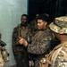 A group of military personnel in two distinct camouflage uniforms are gathered in what appears to be a briefing or coordination session inside a building. Some wear MARPAT (Marine pattern) uniforms while others wear OCP (Operational Camouflage Pattern) uniforms, suggesting a joint or multinational military exercise or operation. One soldier in the center appears to be speaking or gesturing, indicating a leadership or coordination role.