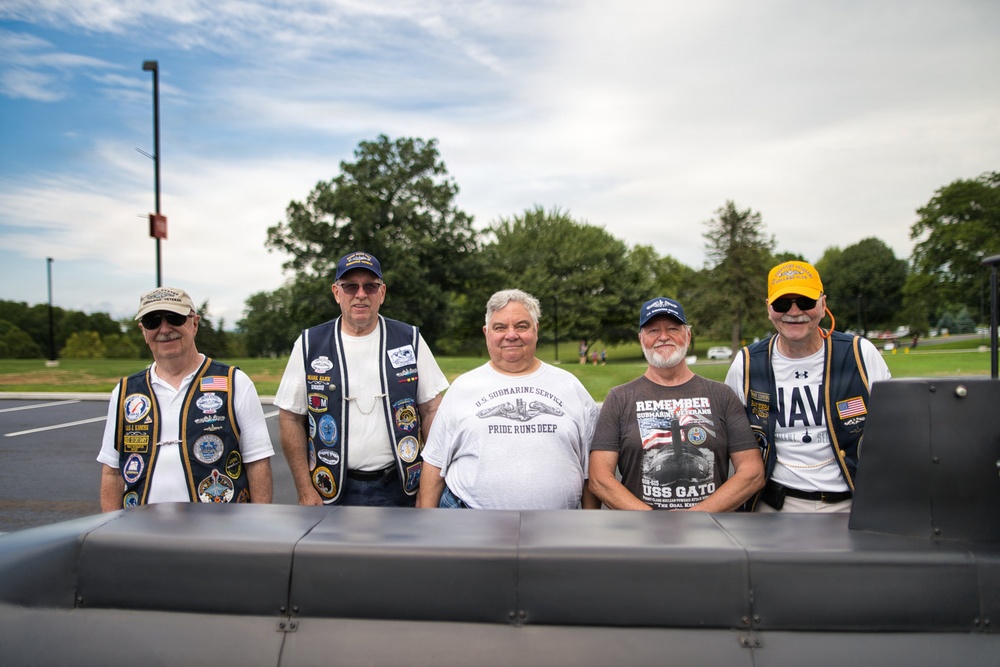 Five older male veterans pose together outdoors, likely at a memorial or veterans event. They are wearing vests and shirts with military patches, insignia, and text referencing U.S. Navy submarine service. A gray metal structure, possibly a submarine replica or monument, is visible in the foreground.