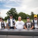 Five older male veterans pose together outdoors, likely at a memorial or veterans event. They are wearing vests and shirts with military patches, insignia, and text referencing U.S. Navy submarine service. A gray metal structure, possibly a submarine replica or monument, is visible in the foreground.