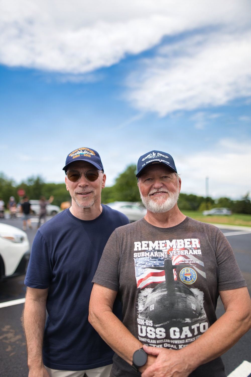 Two older men stand together in a parking lot, both wearing U.S. Navy submarine-related caps and apparel. The man on the right wears a shirt reading 'Remember Submarine Veterans' and 'USS GATO SSN-615 Permit-Class Nuclear-Powered Attack Submarine.' Both appear to be submarine veterans or enthusiasts attending a commemorative or community event.