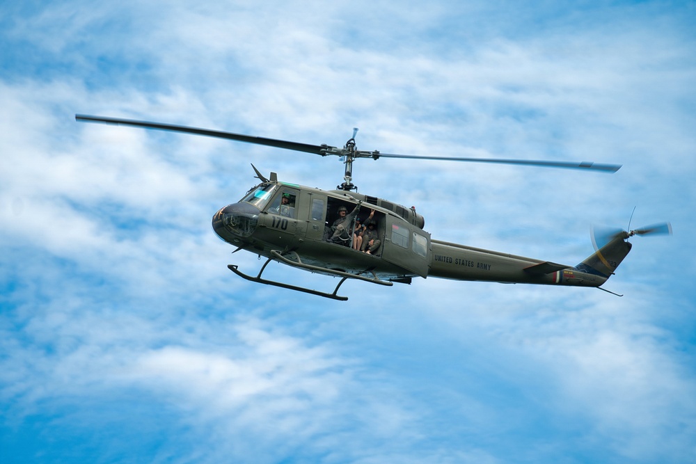 A United States Army UH-1 'Huey' helicopter (tail number 170) is captured in flight against a partly cloudy blue sky. The olive drab military helicopter has its side door open with crew members visible inside. This appears to be either an airshow demonstration or active military flight, likely a restored or preserved Vietnam-era aircraft.