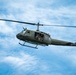 A United States Army UH-1 'Huey' helicopter (tail number 170) is captured in flight against a partly cloudy blue sky. The olive drab military helicopter has its side door open with crew members visible inside. This appears to be either an airshow demonstration or active military flight, likely a restored or preserved Vietnam-era aircraft.