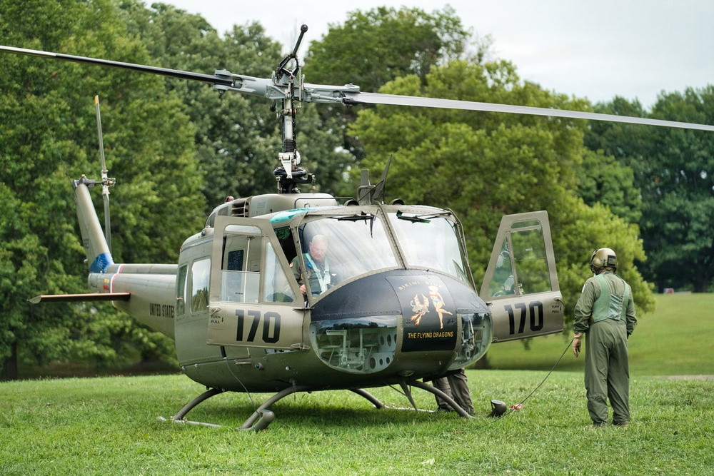 A restored U.S. Army UH-1 'Huey' helicopter marked with the number '170' and 'The Flying Dragons' nose art is parked on a grass field. The aircraft bears 'United States' markings and olive drab paint consistent with Vietnam War-era military livery. Two individuals are present — one seated in the cockpit and one standing nearby in a flight suit and helmet.