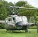 A restored U.S. Army UH-1 'Huey' helicopter marked with the number '170' and 'The Flying Dragons' nose art is parked on a grass field. The aircraft bears 'United States' markings and olive drab paint consistent with Vietnam War-era military livery. Two individuals are present — one seated in the cockpit and one standing nearby in a flight suit and helmet.