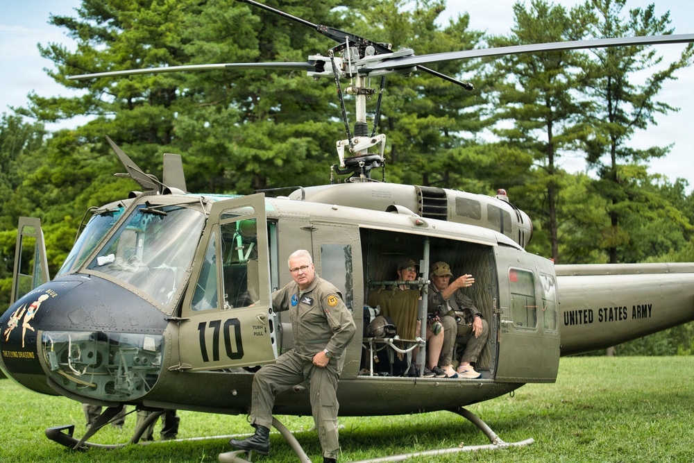 A United States Army UH-1 'Huey' helicopter numbered 170, marked with 'The Flying Dragons' nose art, is displayed on a grass field. A man in an olive drab flight suit leans against the aircraft while two individuals sit in the open cabin doorway. The setting appears to be an outdoor airshow or military heritage event.