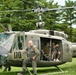 A United States Army UH-1 'Huey' helicopter numbered 170, marked with 'The Flying Dragons' nose art, is displayed on a grass field. A man in an olive drab flight suit leans against the aircraft while two individuals sit in the open cabin doorway. The setting appears to be an outdoor airshow or military heritage event.