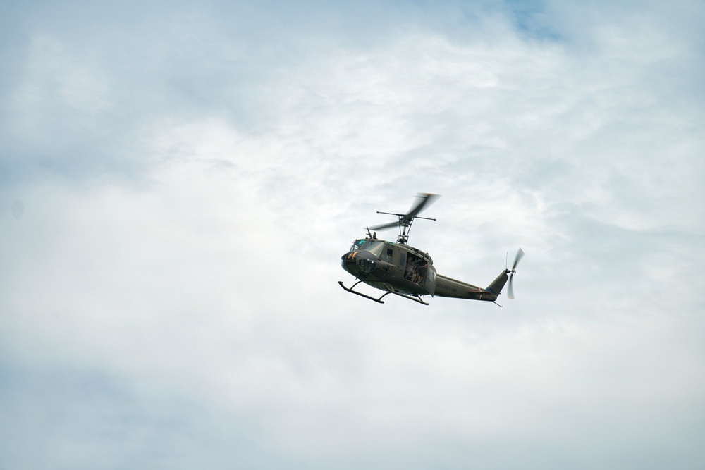 A military utility helicopter, consistent with a Bell UH-1 'Huey' design, is photographed in flight against a partly cloudy sky. The aircraft is painted in olive drab/military green livery and appears to be in level flight. The image is taken from a low angle looking upward, providing a clear view of the helicopter's underside and rotor systems.