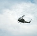 A military utility helicopter, consistent with a Bell UH-1 'Huey' design, is photographed in flight against a partly cloudy sky. The aircraft is painted in olive drab/military green livery and appears to be in level flight. The image is taken from a low angle looking upward, providing a clear view of the helicopter's underside and rotor systems.