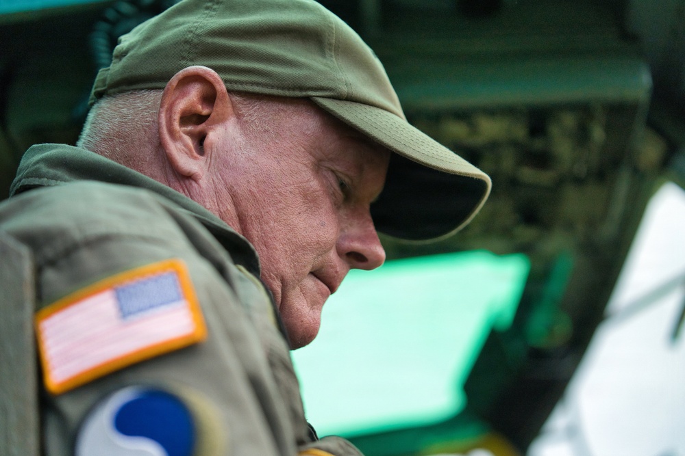 A middle-aged to older male is shown in close profile view, wearing olive/khaki military-style clothing and a matching cap. A US flag patch with an orange border is visible on his left shoulder, along with what appears to be a unit or division patch below it. He appears to be looking downward, possibly reviewing documents or equipment in a military or operational context.