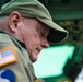 A middle-aged to older male is shown in close profile view, wearing olive/khaki military-style clothing and a matching cap. A US flag patch with an orange border is visible on his left shoulder, along with what appears to be a unit or division patch below it. He appears to be looking downward, possibly reviewing documents or equipment in a military or operational context.