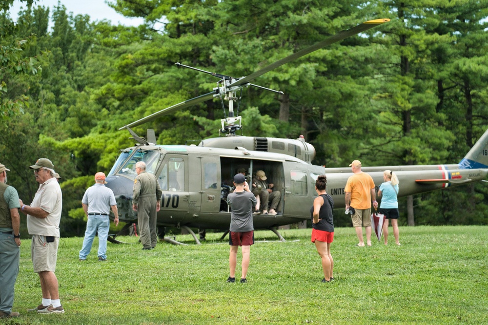A military UH-1 'Huey' helicopter, marked with the number '170' and 'UNITED' (States Army) on its fuselage, is displayed on a grassy field surrounded by civilian visitors. The scene appears to be a public airshow, military heritage event, or museum display where attendees are viewing and photographing the aircraft up close. Several people are gathered around the open cabin of the helicopter, with some appearing to sit inside or inspect the interior.