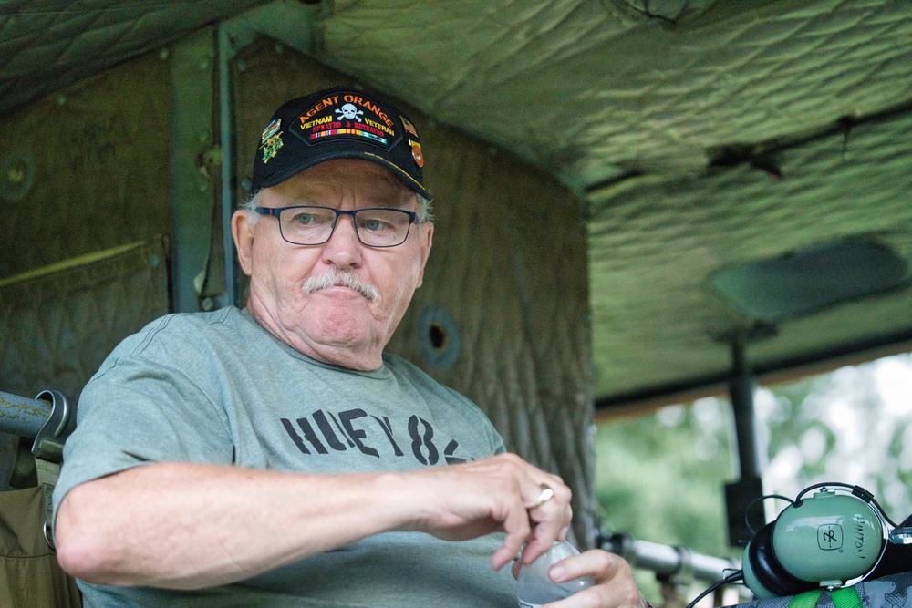 An older man sits inside what appears to be a military vehicle or tent structure. He is wearing a black cap reading 'Agent Orange Vietnam Veteran' with a skull emblem, and a gray t-shirt reading 'HUEY 84'. He appears to be at a military vehicle show or veteran event, holding a small bottle.