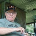 An older man sits inside what appears to be a military vehicle or tent structure. He is wearing a black cap reading 'Agent Orange Vietnam Veteran' with a skull emblem, and a gray t-shirt reading 'HUEY 84'. He appears to be at a military vehicle show or veteran event, holding a small bottle.