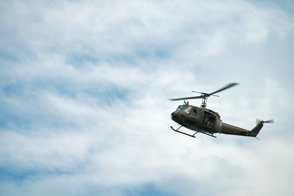 A military utility helicopter, consistent with a Bell UH-1 'Huey' design, is captured in flight against a partly cloudy sky. The aircraft is painted in olive drab/army green livery and appears to have occupants visible through the open side door. The main rotor blades show motion blur, indicating active flight.