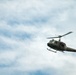 A military utility helicopter, consistent with a Bell UH-1 'Huey' design, is captured in flight against a partly cloudy sky. The aircraft is painted in olive drab/army green livery and appears to have occupants visible through the open side door. The main rotor blades show motion blur, indicating active flight.