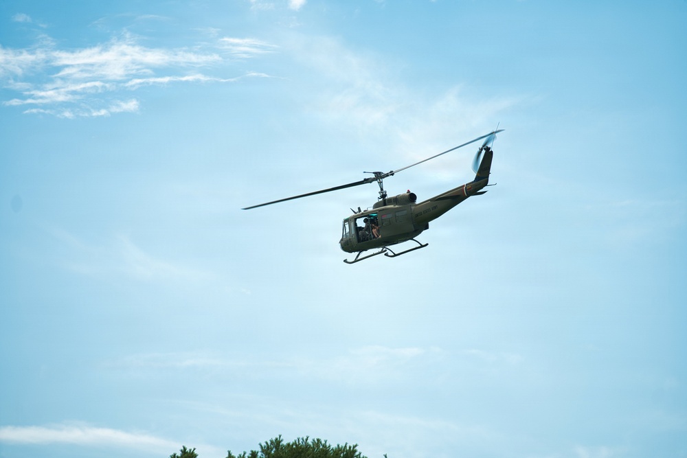 A military-style utility helicopter, consistent with a UH-1 'Huey' design, is photographed in flight against a clear blue sky with light cloud cover. The helicopter is painted in olive drab/army green livery and appears to be flying at low-to-medium altitude. At least one occupant is visible through the open side door of the aircraft.