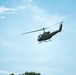 A military-style utility helicopter, consistent with a UH-1 'Huey' design, is photographed in flight against a clear blue sky with light cloud cover. The helicopter is painted in olive drab/army green livery and appears to be flying at low-to-medium altitude. At least one occupant is visible through the open side door of the aircraft.