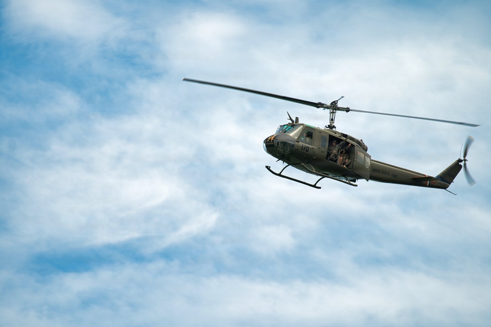 A United States Army olive-drab military helicopter is captured in flight against a partly cloudy blue sky. The aircraft appears to be a Bell UH-1 'Huey', identifiable by its distinctive silhouette, skid landing gear, and single main rotor. Occupants are visible through the open side door of the helicopter.