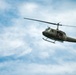 A United States Army olive-drab military helicopter is captured in flight against a partly cloudy blue sky. The aircraft appears to be a Bell UH-1 'Huey', identifiable by its distinctive silhouette, skid landing gear, and single main rotor. Occupants are visible through the open side door of the helicopter.