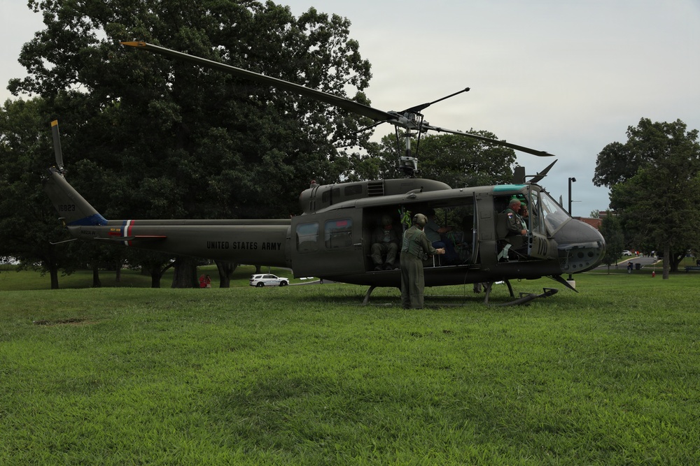 A United States Army UH-1 'Huey' helicopter is landed on a grassy field, with several personnel in flight suits and helmets gathered around and inside the aircraft. The helicopter bears 'UNITED STATES ARMY' markings on its olive drab fuselage along with red, white, and blue insignia. The setting appears to be a park or military installation grounds with large trees and an overcast sky in the background.