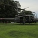 A United States Army UH-1 'Huey' helicopter is landed on a grassy field, with several personnel in flight suits and helmets gathered around and inside the aircraft. The helicopter bears 'UNITED STATES ARMY' markings on its olive drab fuselage along with red, white, and blue insignia. The setting appears to be a park or military installation grounds with large trees and an overcast sky in the background.