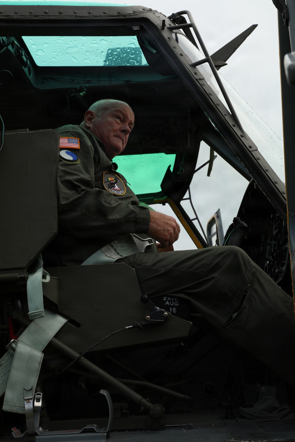An older male in a military flight suit is seated in the cockpit or crew area of what appears to be a military helicopter, likely a UH-1 'Huey' or similar vintage aircraft. He wears an American flag patch, a unit patch, and what appears to be a Vietnam Service ribbon patch on his flight suit. The aircraft door is open and the windows show water droplets, suggesting overcast or rainy conditions.
