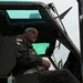 An older male in a military flight suit is seated in the cockpit or crew area of what appears to be a military helicopter, likely a UH-1 'Huey' or similar vintage aircraft. He wears an American flag patch, a unit patch, and what appears to be a Vietnam Service ribbon patch on his flight suit. The aircraft door is open and the windows show water droplets, suggesting overcast or rainy conditions.