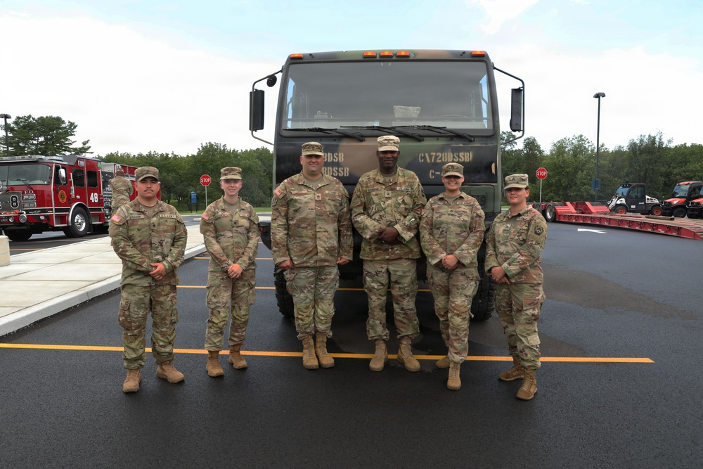 Six U.S. Army soldiers in OCP (Operational Camouflage Pattern) uniforms pose together in front of a large military tactical truck. The group includes both male and female soldiers, standing in a paved area that appears to be a military installation or base. A fire truck marked 'F.D. 48' is visible in the background along with trees and other equipment.
