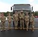 Six U.S. Army soldiers in OCP (Operational Camouflage Pattern) uniforms pose together in front of a large military tactical truck. The group includes both male and female soldiers, standing in a paved area that appears to be a military installation or base. A fire truck marked 'F.D. 48' is visible in the background along with trees and other equipment.