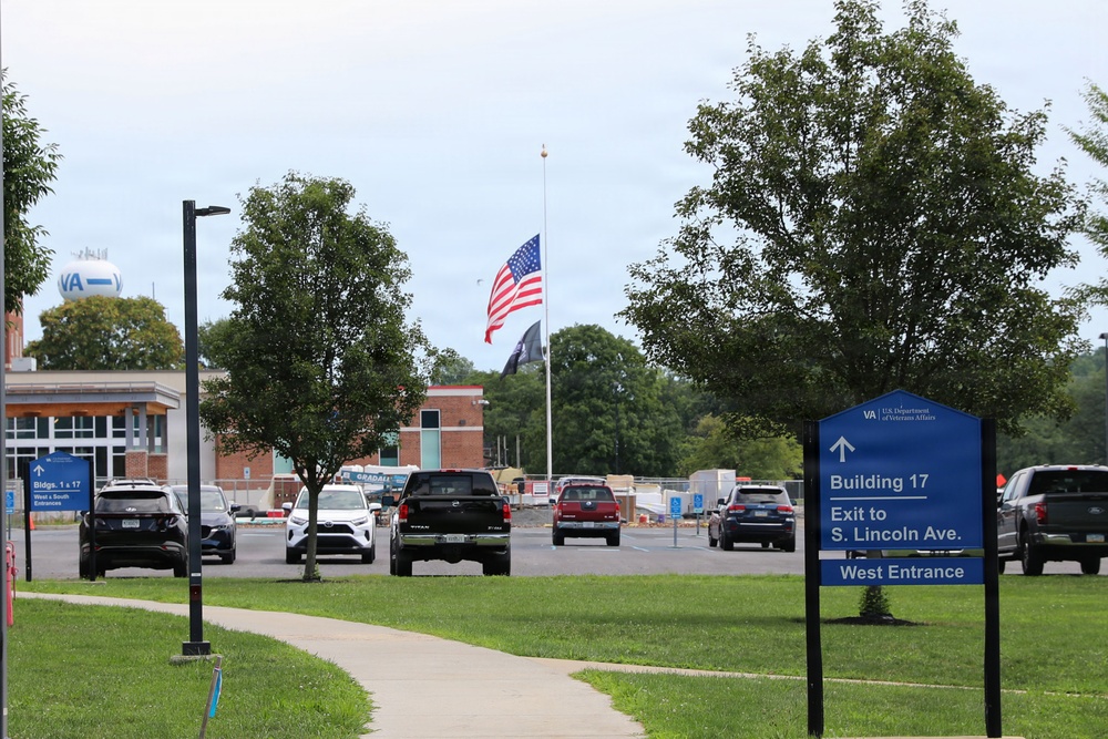 The image shows a U.S. Department of Veterans Affairs (VA) facility campus with a road and parking area in the foreground. An American flag and what appears to be a POW/MIA flag are flying at half-staff on a central flagpole. Directional signage guides visitors toward Building 17, the West Entrance, and an exit to S. Lincoln Ave.