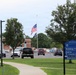 The image shows a U.S. Department of Veterans Affairs (VA) facility campus with a road and parking area in the foreground. An American flag and what appears to be a POW/MIA flag are flying at half-staff on a central flagpole. Directional signage guides visitors toward Building 17, the West Entrance, and an exit to S. Lincoln Ave.