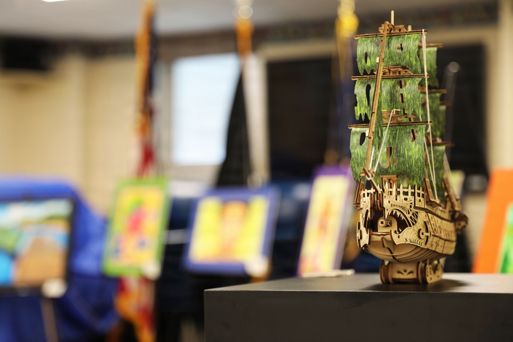 A detailed wooden model sailing ship with green-painted sails is displayed in sharp focus on a dark platform in the foreground. The ship appears to be a laser-cut wooden puzzle model, possibly resembling a fantasy or pirate-style vessel. In the blurred background, colorful artwork pieces are displayed on chairs or easels in what appears to be an indoor exhibition or display event.